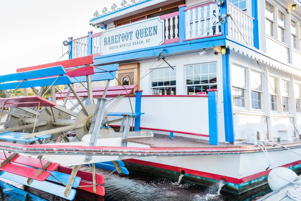 This lively exterior scene features the iconic Barefoot Queen paddlewheel boat docked by a colorful waterfront building. Guests are seen enjoying outdoor dining on the deck, with scenic water views creating an inviting, festive ambiance.