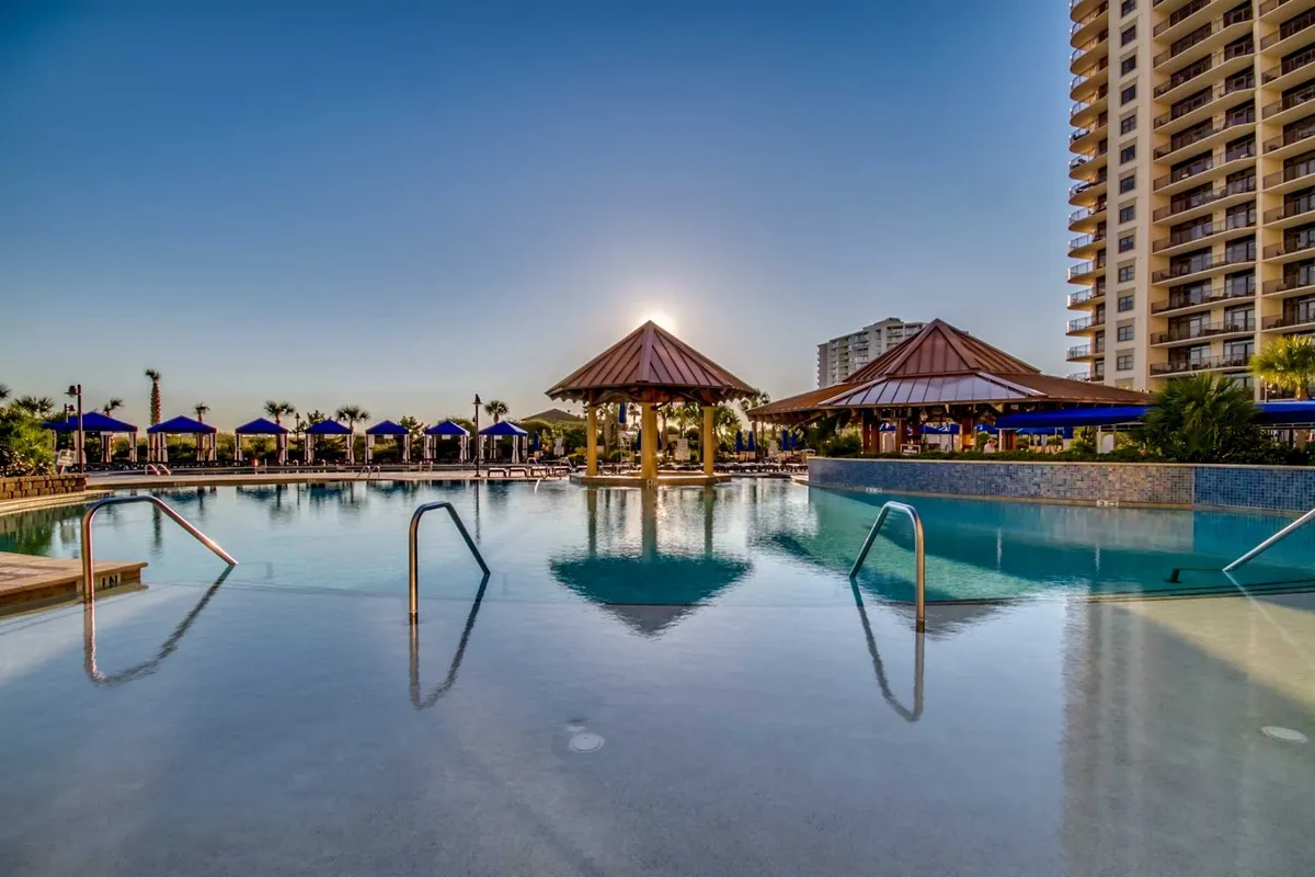 A luxurious pool scene with tropical cabanas, a serene sunset reflecting off the water, and a high-rise building in the background. The image evokes a sense of resort luxury, comfort, and relaxation by the water, ideal for unwinding after a day at the beach.