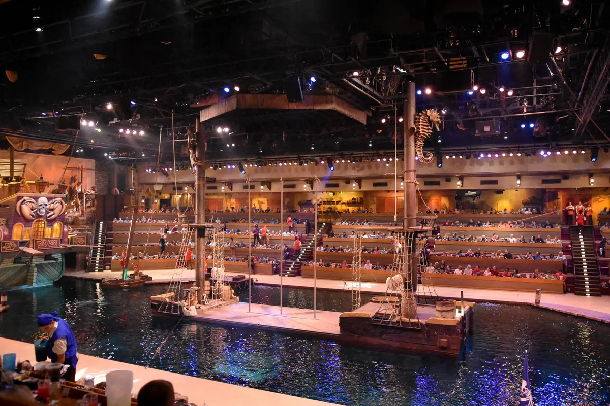 A dramatic interior shot of a themed live performance venue, featuring a large ship structure over water, with audience seating around the stage. The space is immersive and set up for dining and entertainment, suggesting a unique, memorable outing.