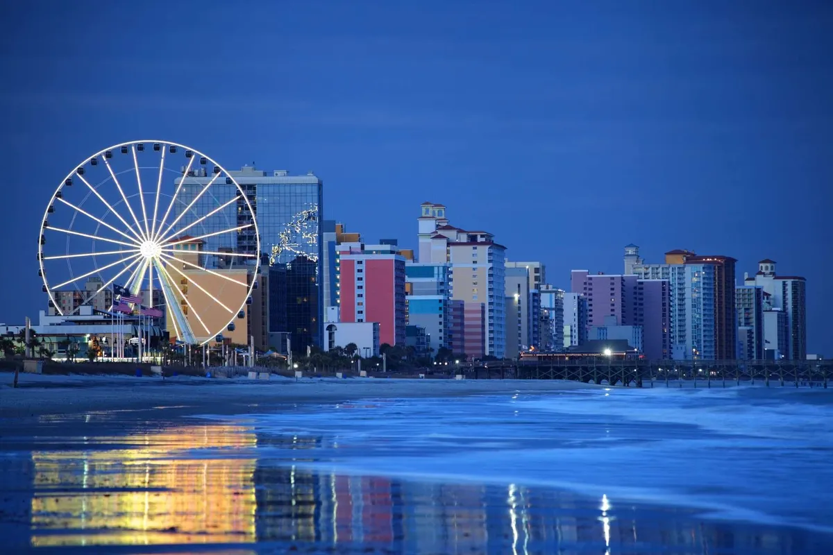 A vibrant evening scene featuring a large ferris wheel and a row of colorful buildings reflecting off the ocean, evoking the lively atmosphere of nearby beachside attractions and nightlife. The view captures the excitement of strolling along the beach at night, with the illuminated skyline and amusement rides in the background.