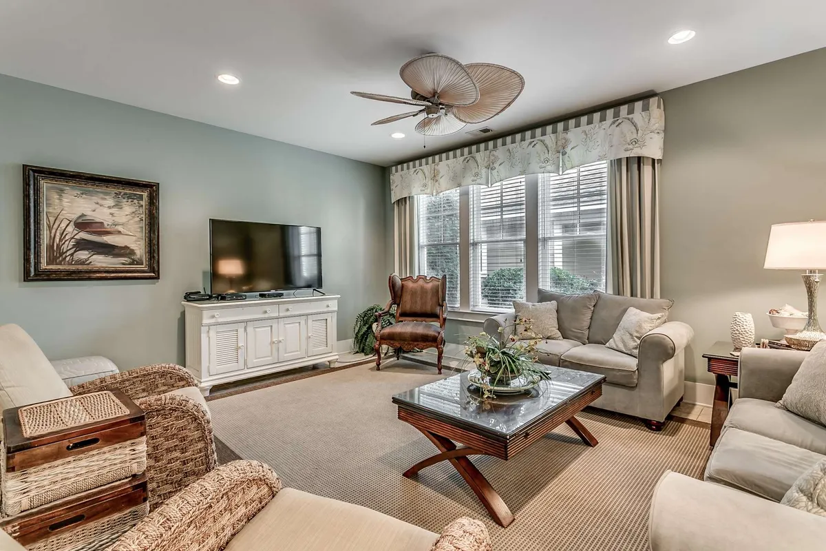 A bright and spacious living room featuring a plush sofa and chair set, a large coffee table, and a prominent television. Ample window seating allows natural light to flood the space, creating a cozy yet open atmosphere ideal for socializing, relaxing, or enjoying entertainment. Subtle coastal decor touches add warmth and a sense of place.