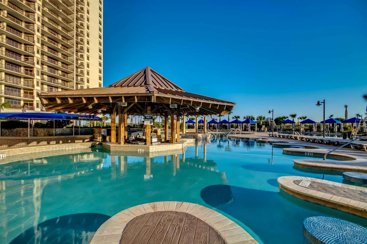A vibrant outdoor pool area surrounded by palm trees, featuring a beautifully maintained pool, pavilion, and plenty of lounge chairs for relaxation. The scene is lively, capturing people swimming, sunbathing, and socializing in a resort-style environment.