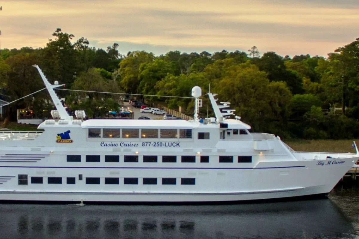 An exterior view of a large white cruise ship surrounded by lush greenery, with prominent casino signage. The ship promises opportunities for cruising, dining, and entertainment, capturing the excitement of signature experiences and exclusive amenities.