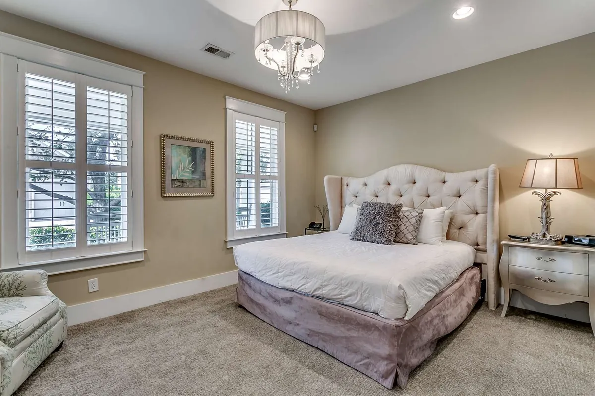 A serene, elegantly decorated bedroom featuring a tufted headboard bed with plush bedding, a cozy armchair by the window, a side table with a lamp, chandelier lighting, and framed artwork. Sunlight streams gently through the window shutters, creating a welcoming retreat ideal for relaxation and quiet time.
