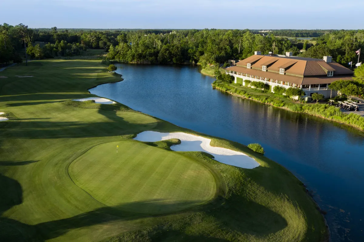 This image features an expansive golf course with a glistening water body and a clubhouse in the background. The lush greens are highlighted by the tranquil lake, and the scene captures people engaged in various activities such as golfing, picnicking, and strolling near the clubhouse.